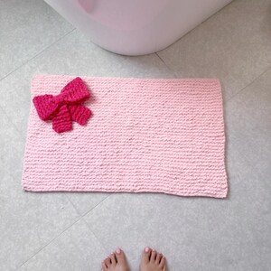 May include: A pink bath mat with a pink crocheted bow on top. The mat is made of a soft, fluffy material and is perfect for adding a touch of style to any bathroom.