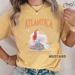 Peut inclure: Un t-shirt jaune d&eacute;lav&eacute; avec une image de dessin anim&eacute; d'Ariel, la Petite Sir&egrave;ne, assise sur un rocher avec les mots "Atlantica" et "Little Mermaid" imprim&eacute;s sur le t-shirt. Le texte sous l'image dit "We have gadgets and gizmos aplenty."