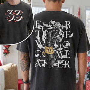 May include: A black t-shirt featuring a graphic on the front with the number 33 surrounded by red and white petals. The back displays a black armored gauntlet holding a rose, wrapped in a tan bandage marked 33, with the text FOR THOSE WHO COME AFTER.