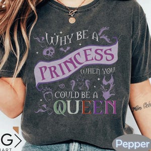 May include: Dark gray t-shirt with the text "Why be a Princess when you could be a Queen". The design includes a purple banner with the word "Princess", and the word "Queen" in multiple colors. The shirt has a vintage, distressed look.