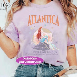 Peut inclure: Un t-shirt violet avec un motif d'Ariel, la Petite Sir&egrave;ne, du film Disney. Le motif comprend le texte "Atlantica" et "Little Mermaid". Le t-shirt a &eacute;galement le texte "Orchid Only With Comfort Color" imprim&eacute; en bas.