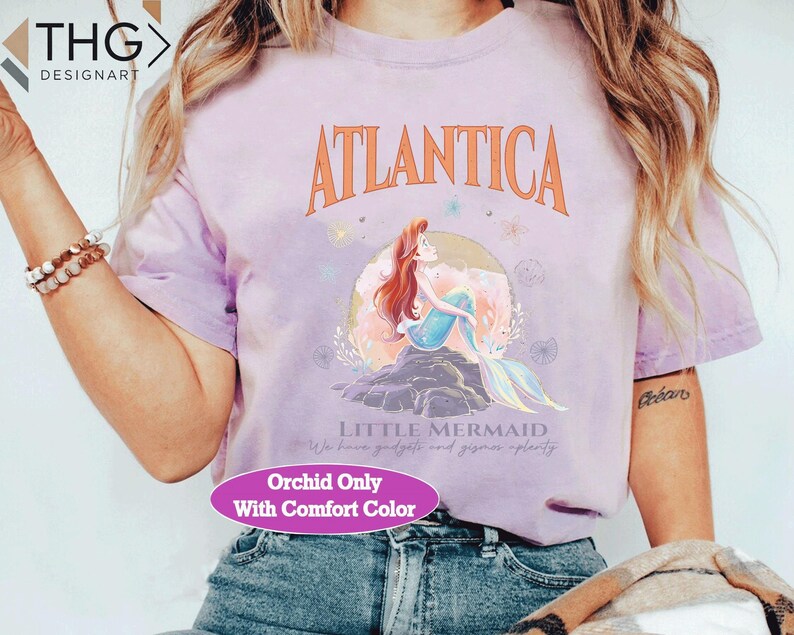 Peut inclure: Un t-shirt violet avec un motif d'Ariel, la Petite Sir&egrave;ne, du film Disney. Le motif comprend le texte "Atlantica" et "Little Mermaid". Le t-shirt a &eacute;galement le texte "Orchid Only With Comfort Color" imprim&eacute; en bas.