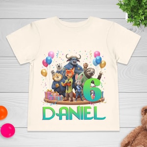 May include: Cream-colored toddler t-shirt featuring a colorful graphic of characters from the animated movie Zootopia, with the number 6 and the name "DANIEL" in green and blue. The design includes balloons and confetti.