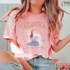 Peut inclure: Un t-shirt rose avec un motif d'Ariel, la Petite Sir&egrave;ne, assise sur un rocher dans l'oc&eacute;an. Le texte "Atlantica" est au-dessus du motif et "Little Mermaid" est en dessous. Le texte "We know gadgets and gizmos aplenty" est en dessous du motif.