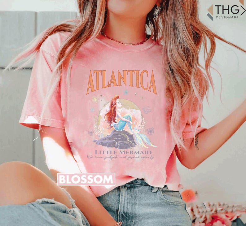 Peut inclure: Un t-shirt rose avec un motif d'Ariel, la Petite Sir&egrave;ne, assise sur un rocher dans l'oc&eacute;an. Le texte "Atlantica" est au-dessus du motif et "Little Mermaid" est en dessous. Le texte "We know gadgets and gizmos aplenty" est en dessous du motif.