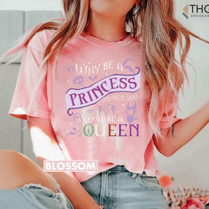 May include: Pink t-shirt with the text "Why be a Princess when you could be a Queen" in purple and white. The shirt has a crew neck and short sleeves. The word "Blossom" is printed on the bottom left.