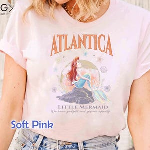 Peut inclure: Un t-shirt rose p&acirc;le avec un motif d'Ariel, la Petite Sir&egrave;ne, assise sur un rocher avec les mots "Atlantica" et "Little Mermaid" imprim&eacute;s sur le t-shirt. Le texte sous le graphique indique "We have gadgets and gizmos aplenty."