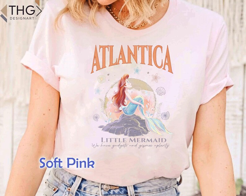 Peut inclure: Un t-shirt rose p&acirc;le avec un motif d'Ariel, la Petite Sir&egrave;ne, assise sur un rocher avec les mots "Atlantica" et "Little Mermaid" imprim&eacute;s sur le t-shirt. Le texte sous le graphique indique "We have gadgets and gizmos aplenty."