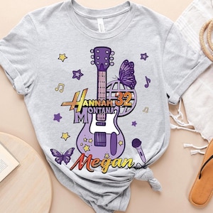 May include: A light gray t-shirt featuring a purple guitar design with the text "Hannah Montana 32 Megan". The design includes butterflies, stars, musical notes, and a microphone. The shirt is tied in a knot at the bottom.