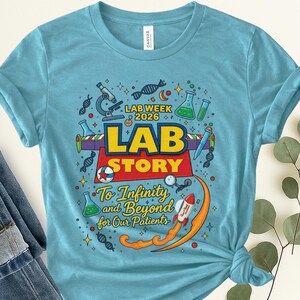 May include: A light blue t-shirt featuring a vibrant graphic. The design incorporates the text "LAB WEEK 2026 LAB STORY To Infinity and Beyond for Our Patients" alongside science-related imagery such as a microscope and DNA strands.