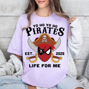 May include: A light purple t-shirt with a pirate-themed graphic. The design features the word "PIRATES" above a pirate hat with a skull and crossbones, a red and black spider-man mask, crossed swords, and the text "EST. 2025 LIFE FOR ME."