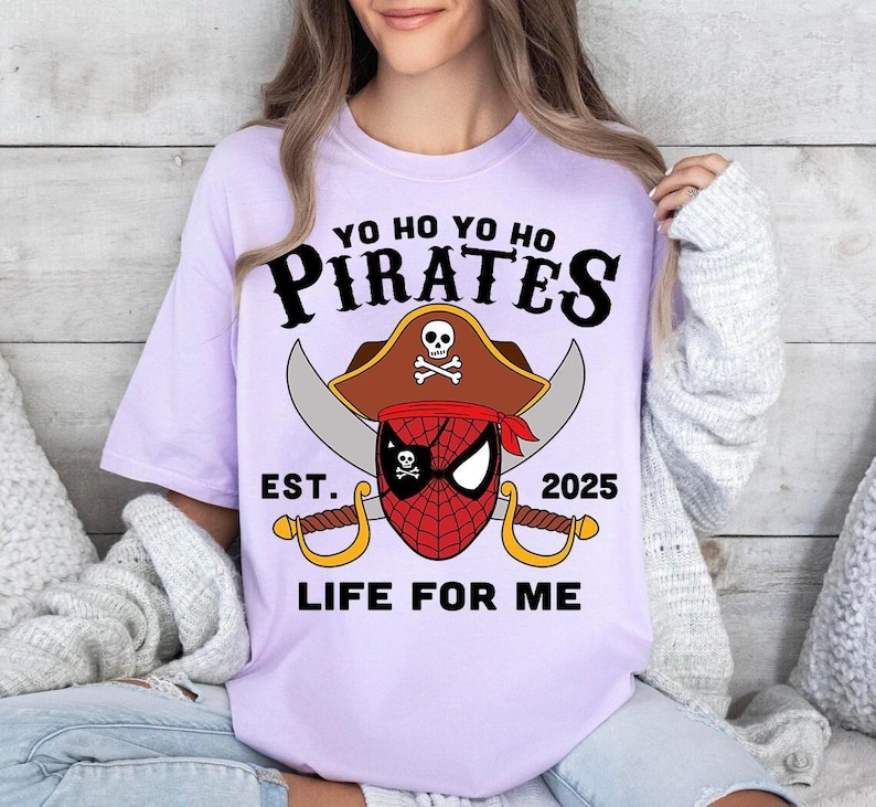 May include: A light purple t-shirt with a pirate-themed graphic. The design features the word "PIRATES" above a pirate hat with a skull and crossbones, a red and black spider-man mask, crossed swords, and the text "EST. 2025 LIFE FOR ME."