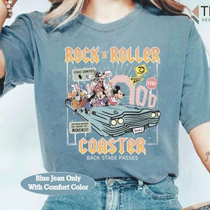 Puede incluir: Una camiseta azul vaquera con un gr&aacute;fico de personajes de Disney en un coche, con el texto "ROCK 'N ROLLER COASTER" en naranja y amarillo. La camiseta tambi&eacute;n tiene el texto "BACK STAGE PASSES" y "Blue Jean Only With Comfort Color".