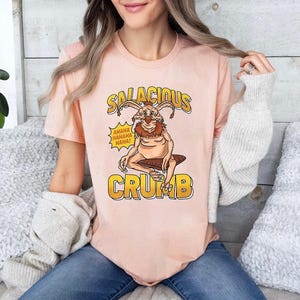 Puede incluir: Camiseta de color melocotón claro con un personaje de dibujos animados y el texto "SALACIOUS CRUMB" en dorado. El personaje tiene orejas grandes y un pecho peludo, sentado en un tronco marrón. Un bocadillo dice "AHAHA HAHAHA HAHA!"