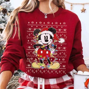 May include: A dark red long-sleeve crewneck sweatshirt featuring a graphic of Mickey Mouse wearing a Santa hat and wrapped in colorful Christmas lights. The background of the graphic includes a white Nordic-style pattern with small Mickey icons.
