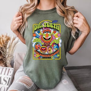 May include: Olive green t-shirt featuring a colorful graphic of Dr. Teeth and The Electric Mayhem. The design includes the band's name, various characters, and the words "World Tour". The shirt has a crew neck and short sleeves.