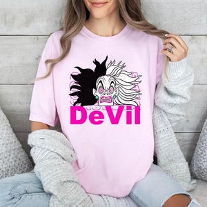 Op de afbeelding: Lichtroze T-shirt met een cartoonillustratie van een schurkenfiguur met zwart en wit haar en een strenge uitdrukking. Het woord "DeVil" staat eronder in felroze.
