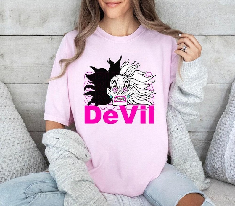 Op de afbeelding: Lichtroze T-shirt met een cartoonillustratie van een schurkenfiguur met zwart en wit haar en een strenge uitdrukking. Het woord "DeVil" staat eronder in felroze.