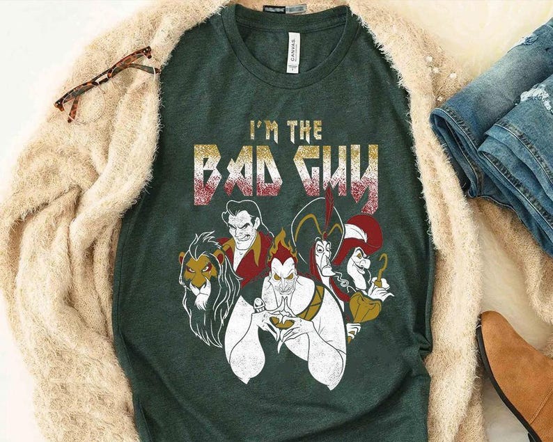 May include: Dark green t-shirt with the text "I'M THE BAD GUY" in gold and red lettering. The shirt features illustrations of several animated villains. The shirt is displayed with a beige cardigan, blue jeans, and brown boots.