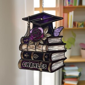 May include: A graduation-themed hanging ornament featuring a stack of books, a mortarboard, a rose, and a butterfly. The books are dark purple with gold accents, and the name "CHARLES" is displayed on the bottom book. The ornament is suspended by a silver chain.