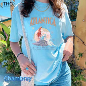 Peut inclure: Un t-shirt bleu clair avec un motif d'Ariel, la Petite Sir&egrave;ne, assise sur un rocher avec les mots "Atlantica" et "Little Mermaid" imprim&eacute;s sur le t-shirt.
