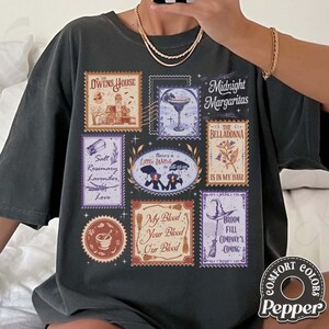 May include: A charcoal gray t-shirt featuring a graphic print of multiple postage stamp-style illustrations. The designs include a house, a cocktail glass, a mortar and pestle, brooms, and text such as Midnight Margaritas and The Owens House.
