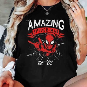 May include: Black t-shirt featuring a red and white graphic of Spider-Man with the text "The Amazing Spider-Man" and "Est. '62". The design includes a spider web and a pointing hand.