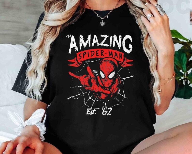 May include: Black t-shirt featuring a red and white graphic of Spider-Man with the text "The Amazing Spider-Man" and "Est. '62". The design includes a spider web and a pointing hand.
