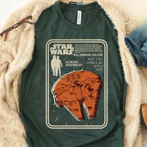 Puede incluir: Camiseta verde oscuro con un diseño de Star Wars que presenta el Halcón Milenario en naranja y blanco. El texto dice "Star Wars Millennium Falcon May the Force Be With You." Parte de una colección de ropa casual.