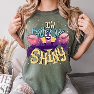May include: Olive green t-shirt featuring a cartoon crab with purple claws and the text "I'd rather be SHINY" in yellow and teal. The crab has a smiling face and is the central design element. The shirt is a casual wear item.