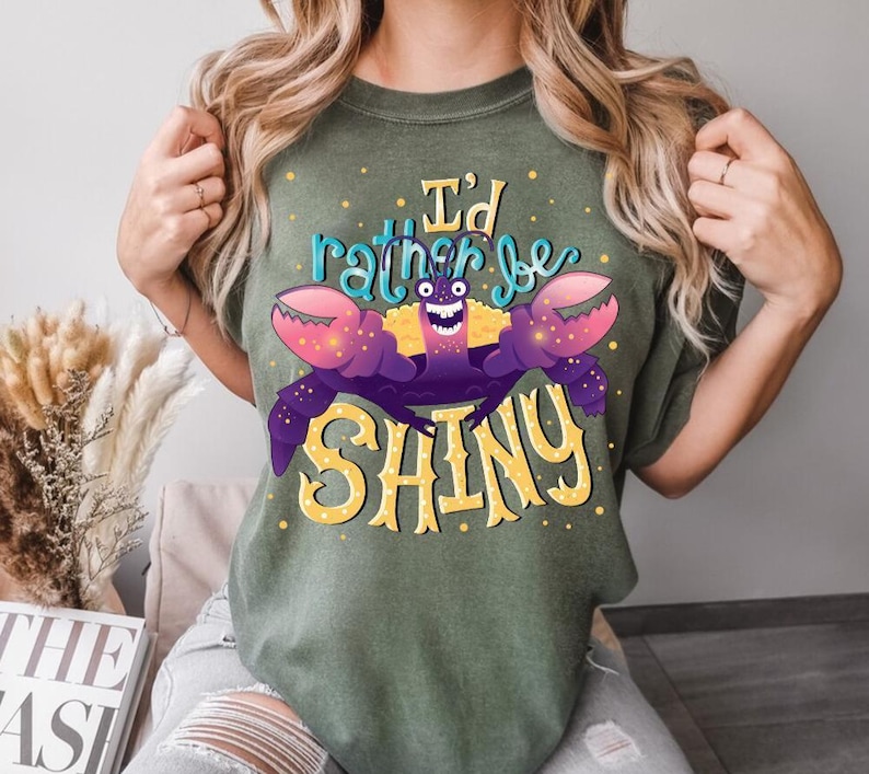 May include: Olive green t-shirt featuring a cartoon crab with purple claws and the text "I'd rather be SHINY" in yellow and teal. The crab has a smiling face and is the central design element. The shirt is a casual wear item.