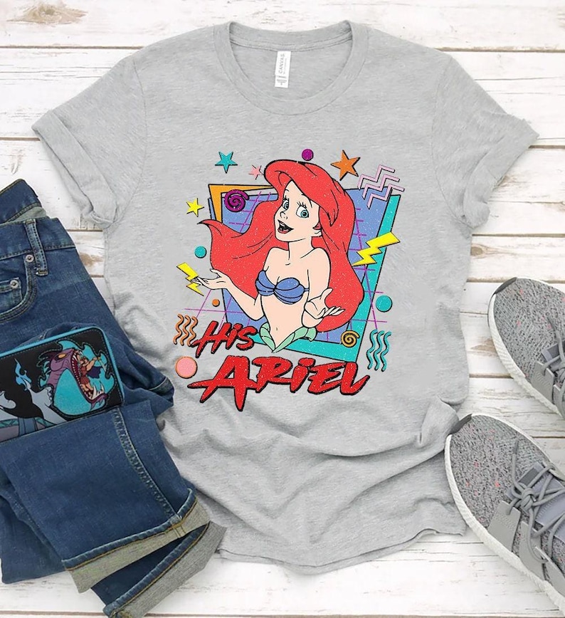 May include: A gray t-shirt featuring a graphic of Ariel from The Little Mermaid. The design includes Ariel with red hair and a blue seashell bra, along with the text "His Ariel" in red. The shirt is paired with blue jeans and gray sneakers.