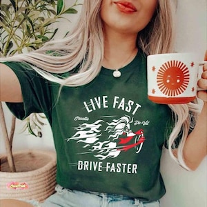 Puede incluir: Camiseta verde oscuro con el texto "LIVE FAST DRIVE FASTER" y un gráfico de Cruella De Vil en una motocicleta. La camiseta también presenta un diseño blanco y rojo. También se ve una taza blanca con un diseño de sol sonriente.