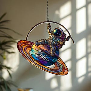 May include: A stained glass suncatcher featuring an astronaut floating above a colorful planet with rings. The astronaut is wearing a detailed spacesuit. The planet has a rainbow color scheme, and the name "John" is written on it in cursive. The suncatcher hangs from a chain.