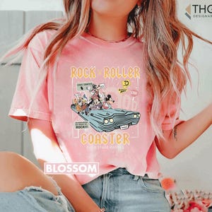 Puede incluir: Camiseta rosa claro con un gr&aacute;fico de personajes de Disney en un coche azul claro, con las palabras "Rock 'n' Roller Coaster" y "Back Stage Passes". La camiseta tambi&eacute;n tiene la palabra "Blossom" impresa.