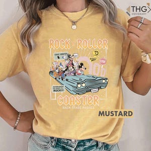 Puede incluir: Camiseta de color mostaza con un gr&aacute;fico de un coche azul claro con personajes de Disney y las palabras "Rock 'n' Roller Coaster". La camiseta tambi&eacute;n tiene el texto "Back Stage Passes" y "Mustard".