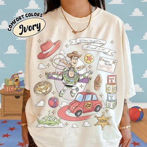 Op de afbeelding: Ivoor kleurig T-shirt met een kleurrijke afbeelding van tekenfilmfiguren. Het ontwerp bevat Woody, Buzz Lightyear, een rode auto en de tekst "You've got a Friend in me."
