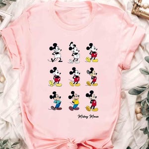 May include: Pink t-shirt featuring a grid of nine Mickey Mouse illustrations in various poses and outfits. The illustrations are in black, white, red, yellow, and blue. The words "Mickey Mouse" are written in cursive at the bottom.