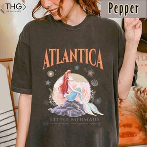 Peut inclure: Un t-shirt noir avec un imprim&eacute; vintage d'Ariel, la Petite Sir&egrave;ne, assise sur un rocher avec les mots "Atlantica" et "Little Mermaid" imprim&eacute;s sur le t-shirt. Le graphique comprend &eacute;galement des coquillages, des &eacute;toiles de mer et des algues.