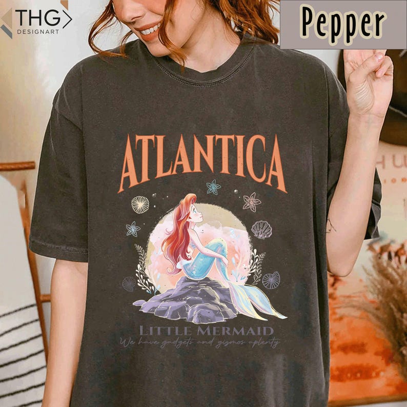 Peut inclure: Un t-shirt noir avec un imprim&eacute; vintage d'Ariel, la Petite Sir&egrave;ne, assise sur un rocher avec les mots "Atlantica" et "Little Mermaid" imprim&eacute;s sur le t-shirt. Le graphique comprend &eacute;galement des coquillages, des &eacute;toiles de mer et des algues.