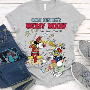 May include: A gray t-shirt featuring a colorful graphic of Mickey Mouse conducting a band, with Donald Duck playing a trumpet. The text "Walt Disney's Mickey Mouse" is above the image, with "The Band Concert" below.
