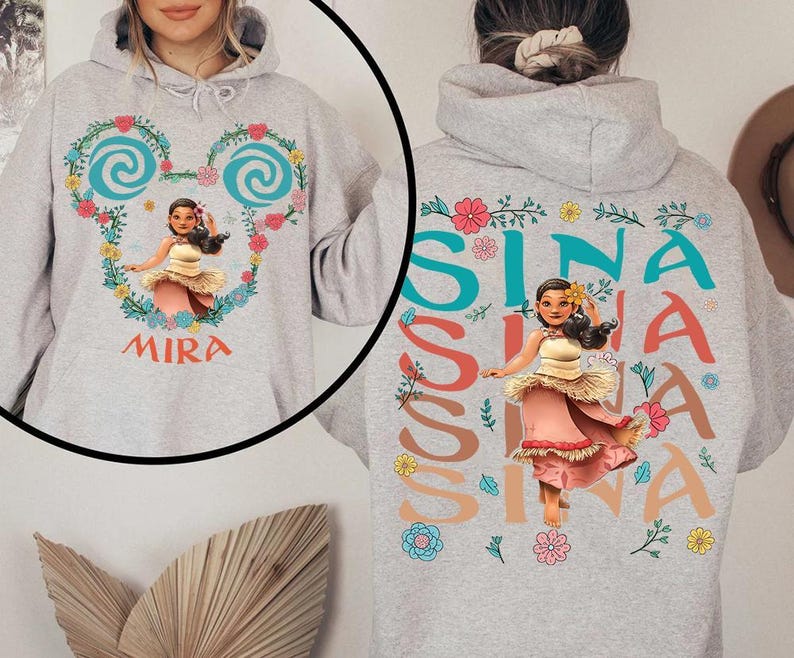 May include: A light grey hoodie with a cartoon character in a pink and cream dress. The front features the name "MIRA" and a heart-shaped floral design. The back of the hoodie displays the name "SINA" in colourful letters.