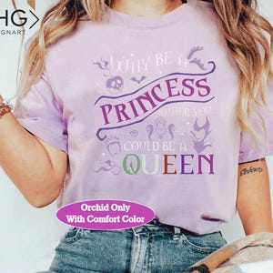 May include: A light purple t-shirt with the text "WHY BE A PRINCESS WHEN YOU COULD BE A QUEEN" in purple, green, and red. The shirt has a crew neck and short sleeves. The text "Orchid Only With Comfort Color" is also visible.