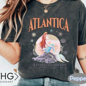 Peut inclure: Un t-shirt gris fonc&eacute; &agrave; l'aspect vieilli avec un motif d'Ariel, la Petite Sir&egrave;ne, avec le texte "Atlantica" et "Little Mermaid" imprim&eacute; sur le devant.