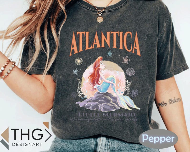 Peut inclure: Un t-shirt gris fonc&eacute; &agrave; l'aspect vieilli avec un motif d'Ariel, la Petite Sir&egrave;ne, avec le texte "Atlantica" et "Little Mermaid" imprim&eacute; sur le devant.