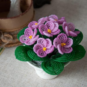 Puede incluir: Un arreglo floral de cuentas en una maceta blanca. Las flores son de color rosa con centros amarillos y hojas verdes. Las flores están hechas de cuentas y están dispuestas en un ramo.