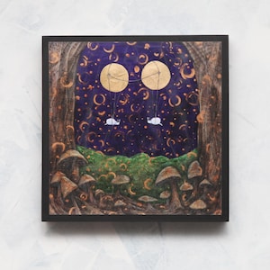May include: Square framed art print featuring a whimsical forest scene. Two white creatures float beneath two golden orbs in a starry night sky. The forest floor is populated with mushrooms and green foliage. The artwork is framed in a black square.