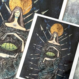 May include: Two art prints featuring a watercolor illustration of a figure with long brown hair, a golden halo, and a dark cloak. The figure holds a green oval object. The background is dark blue with arrows and a golden moon.