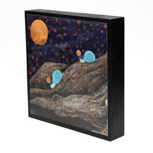 May include: A framed art piece featuring a whimsical landscape with two light blue snails. The background is a dark blue night sky with a large orange moon and scattered golden orbs. The artwork is set in a black frame.