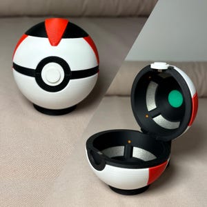 May include: A white, black, and red spherical container, resembling a ball, with a circular button on the front. The container is shown both closed and open, revealing a black interior with a green circle.